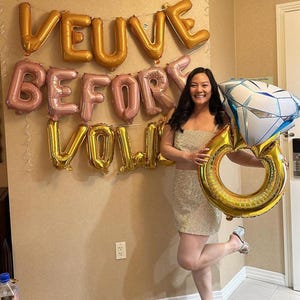 May include: A woman in a sparkly gold dress holds a large gold ring balloon and a diamond balloon. The balloons spell out "Veuve Before You" on the wall behind her.
