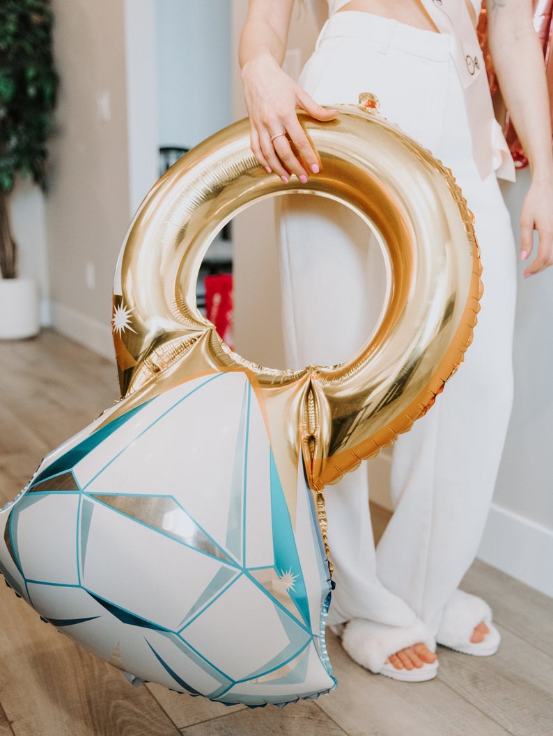 May include: A large, gold ring-shaped balloon with a diamond-shaped base. The diamond base is white with blue and silver geometric designs. The balloon is held by a person wearing white pants and fuzzy slippers.