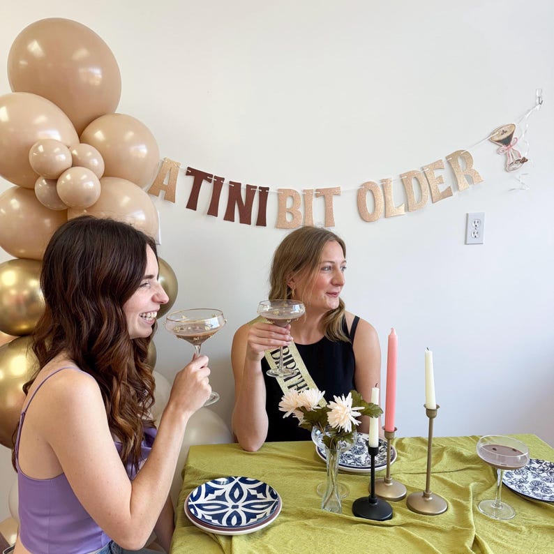 May include: Two women celebrating with cocktails, seated at a table with a green tablecloth. A banner that reads "A TINI BIT OLDER" hangs above them. Balloons in shades of gold and brown are in the background. The table is set with plates, candles, and flowers.