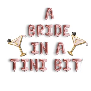 Può includere: Lettere di palloncini in lamina color oro rosa che compongono la frase "A BRIDE IN A TINI BIT" con due palloncini a forma di bicchiere da martini. I palloncini sono disposti su sfondo bianco, perfetti per un addio al nubilato.
