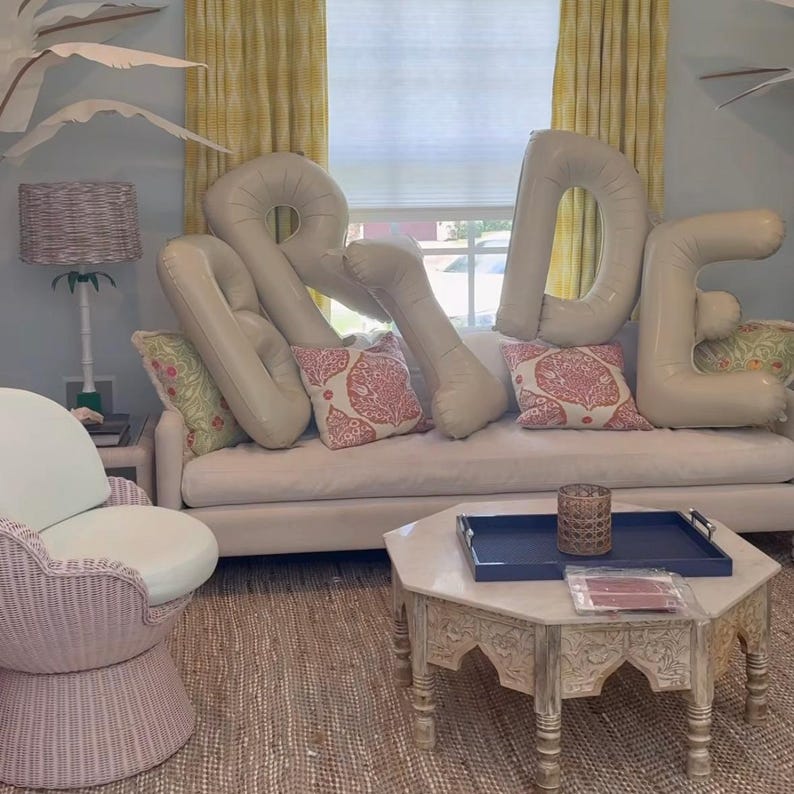 Peut inclure: Grands ballons gonflables beiges formant le mot BRIDE dispos&eacute;s sur un canap&eacute; clair avec des coussins &agrave; motifs. La sc&egrave;ne comprend une chaise en osier rose p&acirc;le, une table basse en bois avec un plateau bleu et une lampe en osier.