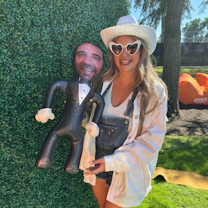 Puede incluir: Una mujer con una camisa blanca y un mono está sosteniendo un hombre inflable grande que lleva un esmoquin. El hombre tiene una cara impresa y una corbata de moño. La mujer lleva un sombrero blanco y gafas de sol en forma de corazón.