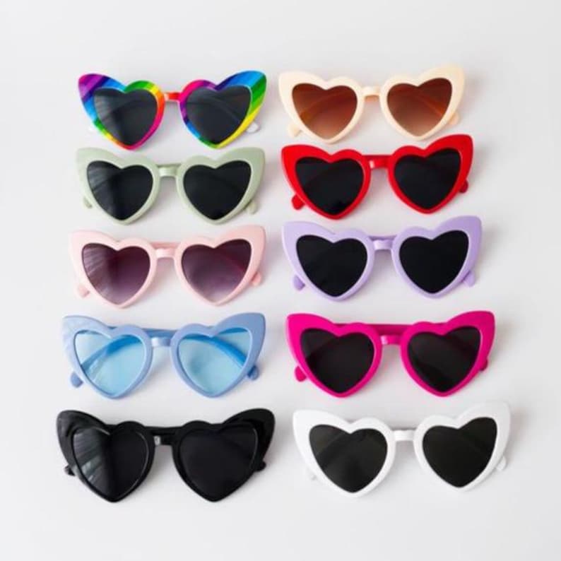 May include: A collection of heart-shaped sunglasses in various colors, including rainbow, cream, red, pink, purple, blue, black, and white. Each pair has dark lenses. The sunglasses are arranged in rows on a white surface.
