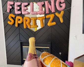 May include: Pink and gold foil balloons spell out "FEELING SPRITZY" above a fireplace. A champagne bottle and orange slice balloon are in the foreground. The champagne bottle has a pink label with gold accents. The orange slice balloon is orange and white.