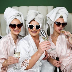 May include: Three people wearing heart-shaped sunglasses and white bath towels on their heads, holding champagne flutes. They are wearing satin robes in shades of pink and white. One person is holding a champagne bottle.