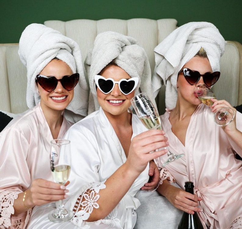 May include: Three people wearing heart-shaped sunglasses and white bath towels on their heads, holding champagne flutes. They are wearing satin robes in shades of pink and white. One person is holding a champagne bottle.