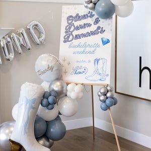 May include: A party scene with balloons and a sign. The sign reads "Olivia's Denim & Diamonds bachelorette weekend" with a drawing of a cowboy boot. Balloons are white, silver, and blue. A large boot-shaped balloon is also present.