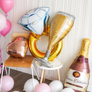 May include: A collection of celebratory balloons, including a diamond ring, champagne glass, and champagne bottle. Pink and white balloons are scattered around the scene, creating a festive atmosphere. The champagne bottle reads "Ch&acirc;teau C&eacute;l&eacute;bration".