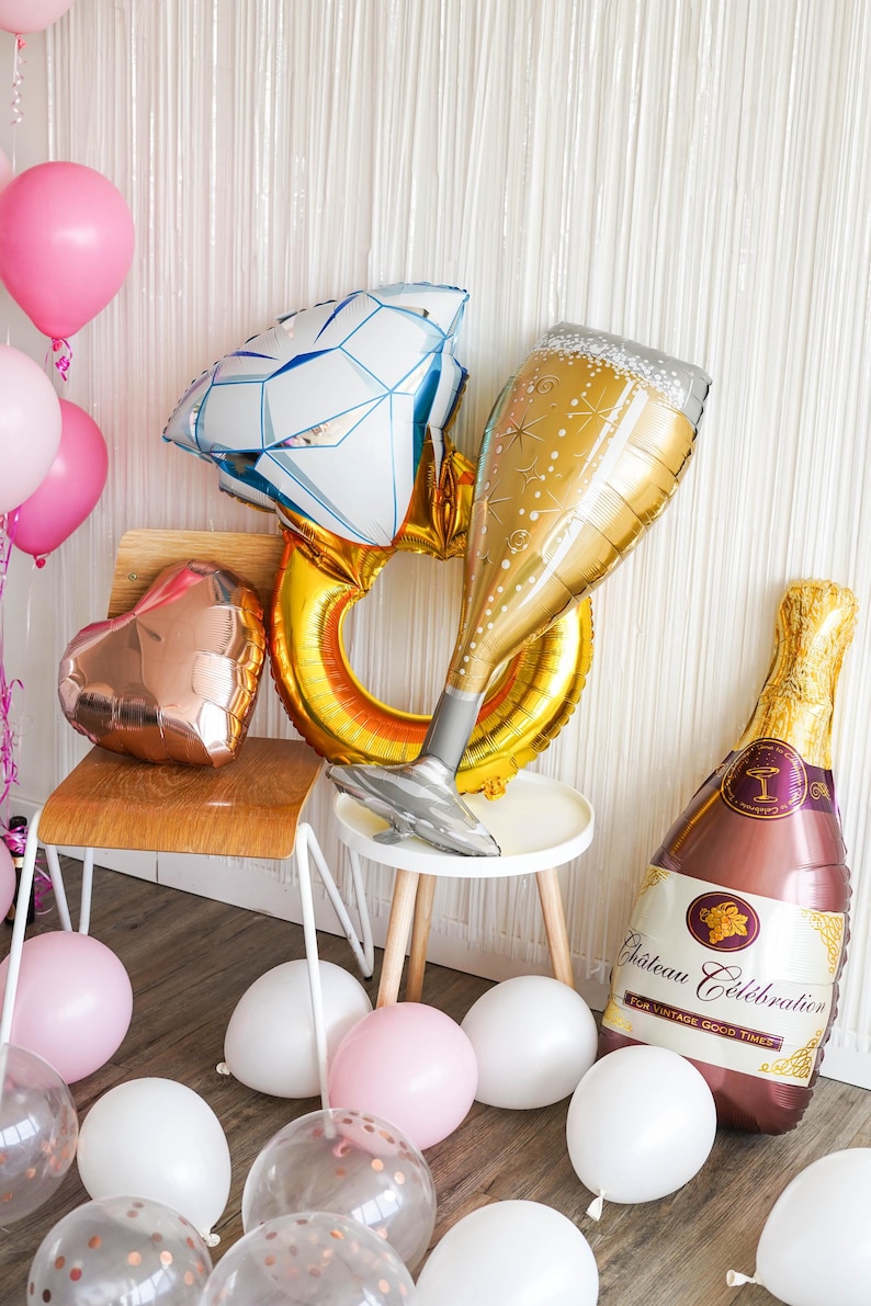 May include: A collection of celebratory balloons, including a diamond ring, champagne glass, and champagne bottle. Pink and white balloons are scattered around the scene, creating a festive atmosphere. The champagne bottle reads "Ch&acirc;teau C&eacute;l&eacute;bration".