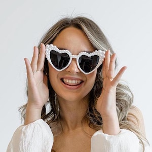 Op de afbeelding: Witte hartvormige zonnebril met zwarte glazen en parelversiering. De zonnebril wordt gedragen door een persoon met lang blond haar.