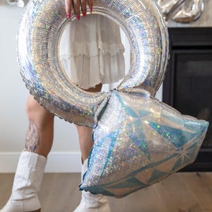 May include: A large, silver ring-shaped balloon with a diamond-shaped accent. The balloon has a holographic, iridescent finish. The diamond-shaped accent is blue and silver. The balloon is held by a person wearing a white dress and boots.