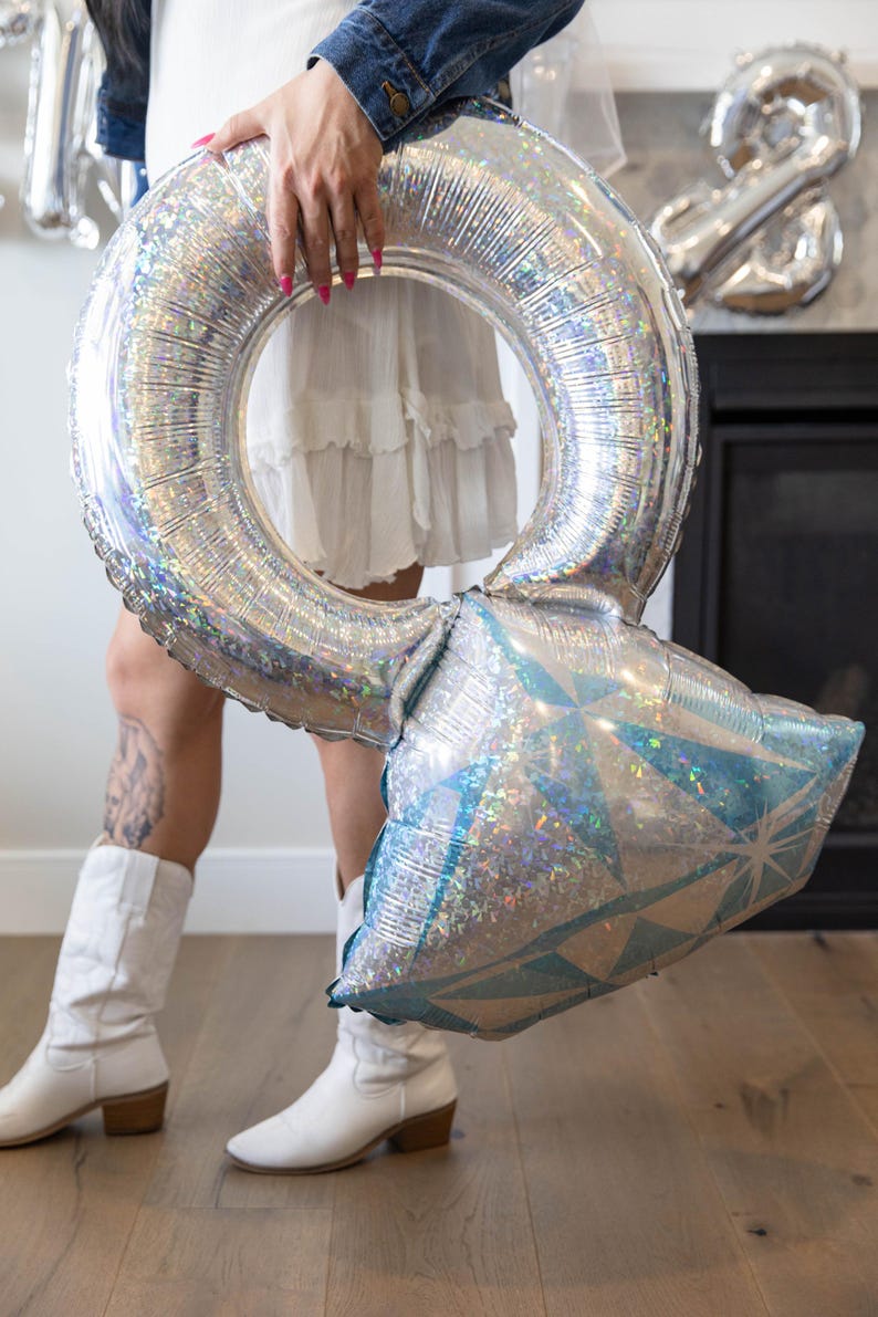 May include: A large, silver ring-shaped balloon with a diamond-shaped accent. The balloon has a holographic, iridescent finish. The diamond-shaped accent is blue and silver. The balloon is held by a person wearing a white dress and boots.