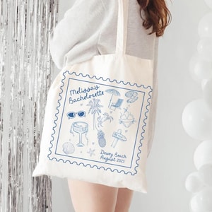 Puede incluir: Un bolso tote de color crema con un diseño azul en forma de sello. El diseño incluye ilustraciones de playa y el texto "Melissa's Bachelorette" y "Dewey Beach August 2025". El bolso tiene asas largas y lo lleva una persona.