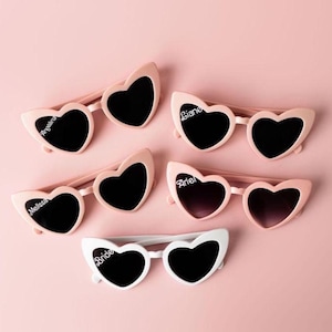 May include: Pink heart-shaped sunglasses with black lenses, some personalized with names like "Angelina" and "Melissa." One pair is white with "Bride" printed on the frame. The sunglasses are arranged on a pink background.