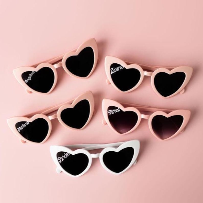 May include: Pink heart-shaped sunglasses with black lenses, some personalized with names like "Angelina" and "Melissa." One pair is white with "Bride" printed on the frame. The sunglasses are arranged on a pink background.