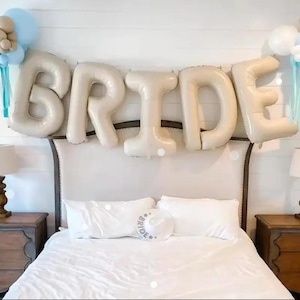 Peut inclure: Un ensemble de grands ballons en aluminium beige formant le mot BRIDE, suspendus au-dessus d'un lit. La d&eacute;coration comprend des grappes de ballons bleu clair et beige sur les c&ocirc;t&eacute;s et un chapeau blanc avec le mot BRIDE imprim&eacute; devant.