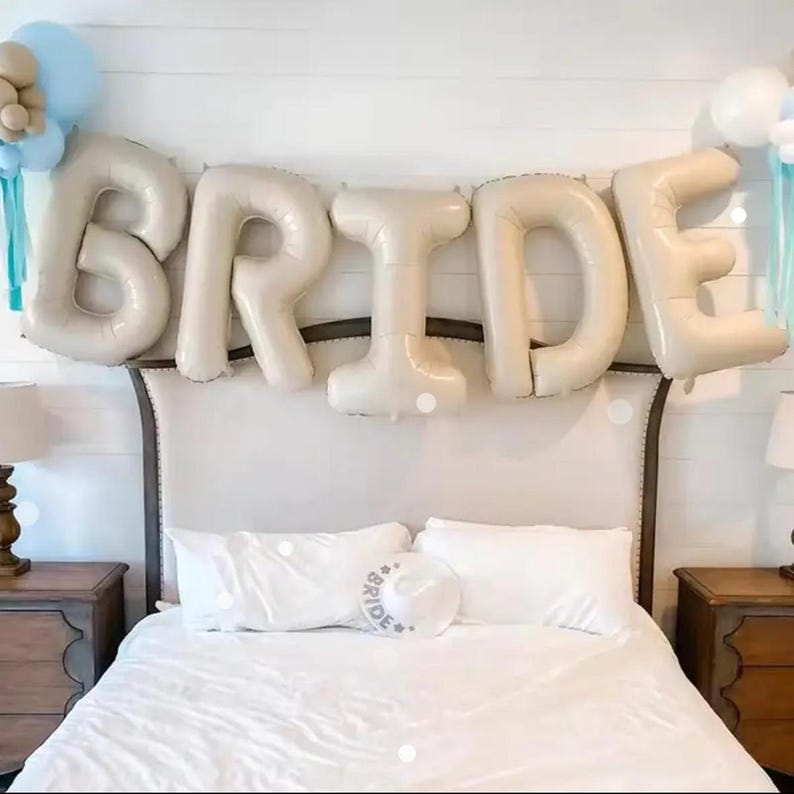 Peut inclure: Un ensemble de grands ballons en aluminium beige formant le mot BRIDE, suspendus au-dessus d'un lit. La d&eacute;coration comprend des grappes de ballons bleu clair et beige sur les c&ocirc;t&eacute;s et un chapeau blanc avec le mot BRIDE imprim&eacute; devant.