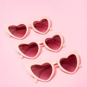 May include: Four pairs of heart-shaped sunglasses. The frames are a pale pink, and the lenses are a darker shade of pink. The sunglasses are arranged diagonally on a solid pink background.