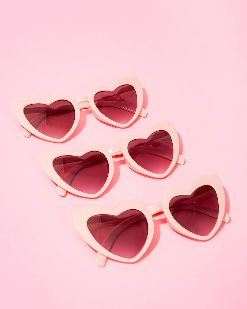 May include: Four pairs of heart-shaped sunglasses. The frames are a pale pink, and the lenses are a darker shade of pink. The sunglasses are arranged diagonally on a solid pink background.
