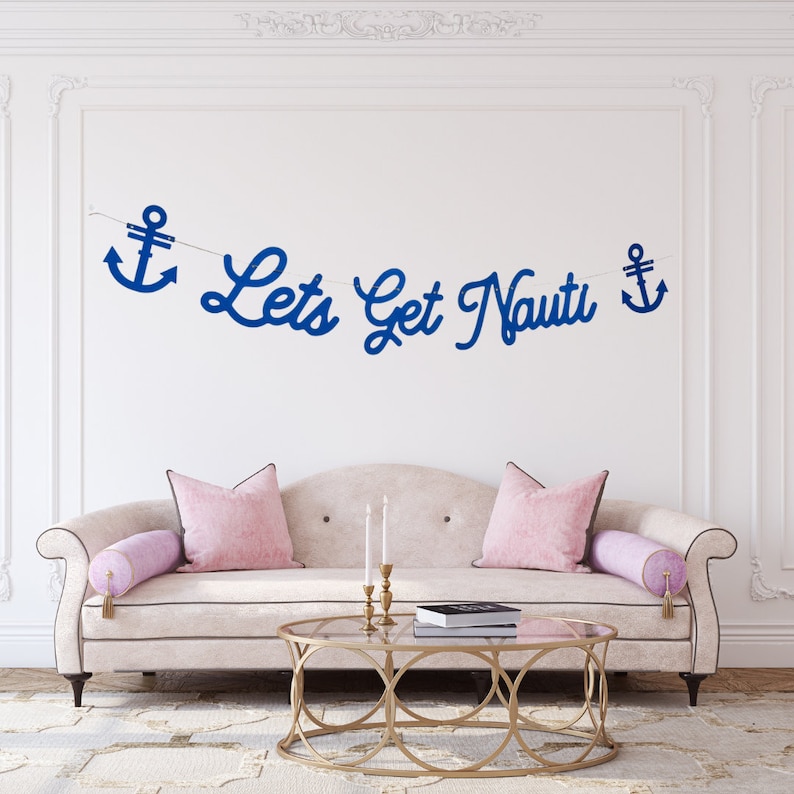 Puede incluir: Un sof&aacute; blanco con cojines rosas y una mesa de centro de metal dorado. Un banner azul con anclas y el texto "Let's Get Nauti" cuelga en la pared detr&aacute;s del sof&aacute;.