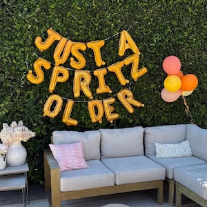 May include: A party scene with a green hedge backdrop. Gold foil balloons spell out "JUST A SPRITZ OLDER." A gray sectional sofa with pink and patterned pillows is in front. A small bouquet of flowers and a cluster of balloons in shades of pink, orange, and yellow complete the scene.