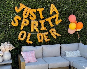 May include: A party scene with a green hedge backdrop. Gold foil balloons spell out "JUST A SPRITZ OLDER." A gray sectional sofa with pink and patterned pillows is in front. A small bouquet of flowers and a cluster of balloons in shades of pink, orange, and yellow complete the scene.