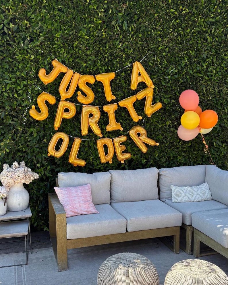 May include: A party scene with a green hedge backdrop. Gold foil balloons spell out "JUST A SPRITZ OLDER." A gray sectional sofa with pink and patterned pillows is in front. A small bouquet of flowers and a cluster of balloons in shades of pink, orange, and yellow complete the scene.