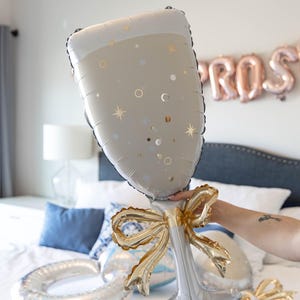 May include: A large champagne glass-shaped balloon in silver and white, decorated with gold stars and dots. The balloon has a gold bow at the base. Other balloons are visible on a bed, with a "PROS" balloon banner in the background.