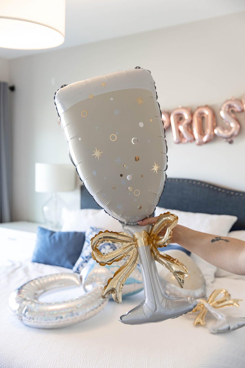 May include: A large champagne glass-shaped balloon in silver and white, decorated with gold stars and dots. The balloon has a gold bow at the base. Other balloons are visible on a bed, with a "PROS" balloon banner in the background.