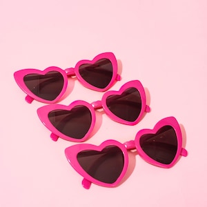 May include: Five pairs of bright pink heart-shaped sunglasses with dark lenses are arranged on a light pink background. The sunglasses have a glossy finish and a playful, romantic aesthetic, perfect for a fun, fashionable look.