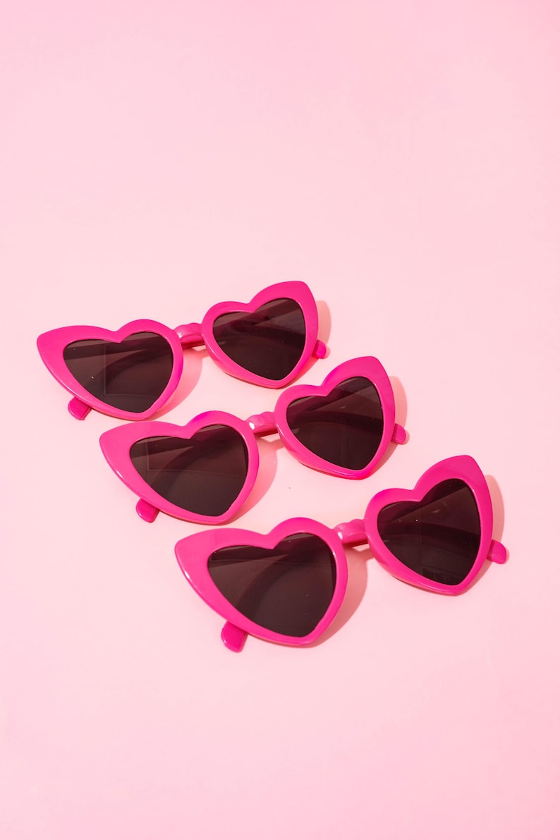 May include: Five pairs of bright pink heart-shaped sunglasses with dark lenses are arranged on a light pink background. The sunglasses have a glossy finish and a playful, romantic aesthetic, perfect for a fun, fashionable look.