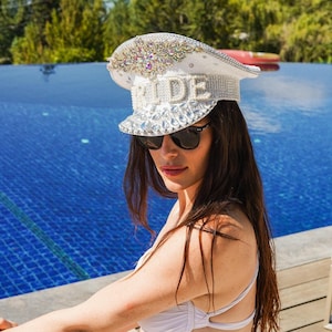 May include: White captain's hat with the word "RIDE" spelled out in silver letters. The hat is embellished with rhinestones and other decorative elements. The person in the image is wearing sunglasses and a white top.