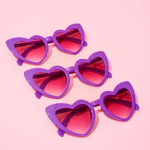 May include: Four pairs of heart-shaped sunglasses with purple frames and gradient pink lenses are arranged on a pink background. The sunglasses have a retro, playful design, perfect for adding a touch of fun to any outfit.