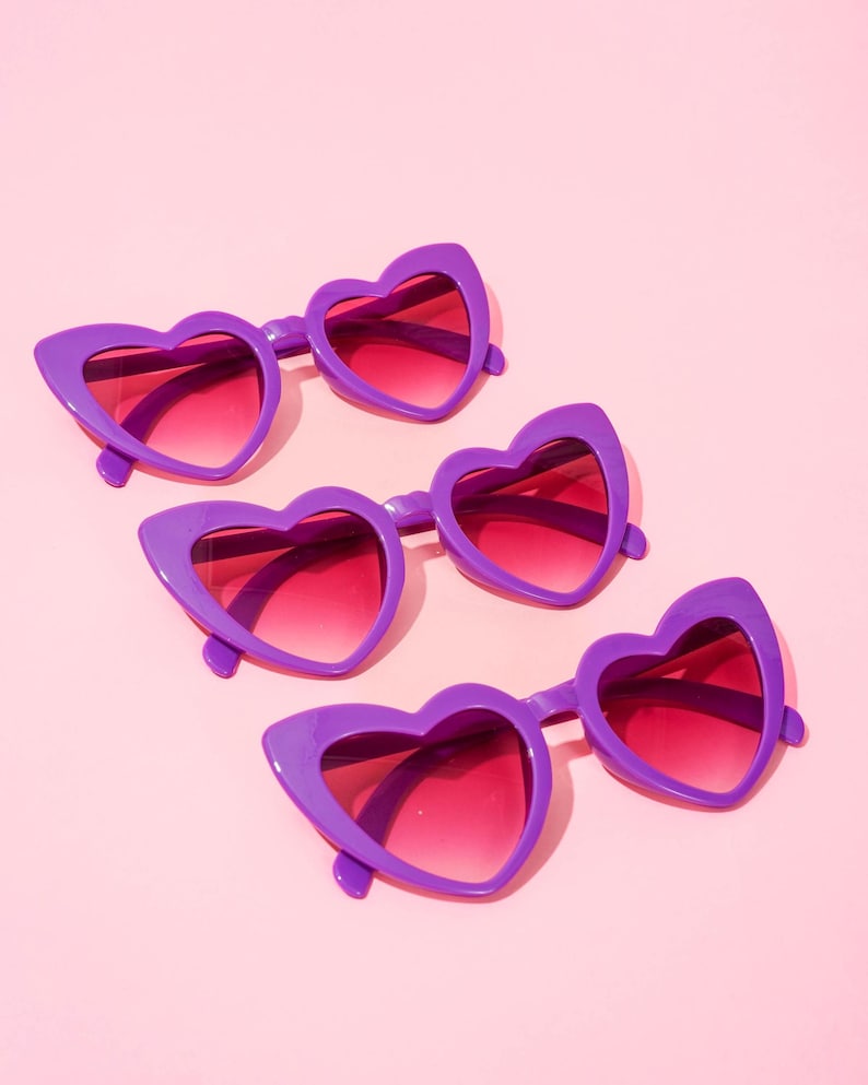 May include: Four pairs of heart-shaped sunglasses with purple frames and gradient pink lenses are arranged on a pink background. The sunglasses have a retro, playful design, perfect for adding a touch of fun to any outfit.