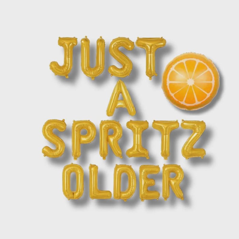 May include: Gold foil balloons spelling out "JUST A SPRITZ OLDER" with an orange slice balloon. The balloons are arranged on a white background. The orange slice balloon is orange and white.