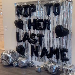 May include: A silver metallic fringe curtain backdrop decorated with black letter balloons spelling out RIP TO HER LAST NAME, accompanied by black heart-shaped balloons and several silver mirrored disco ball decorations placed on the floor.