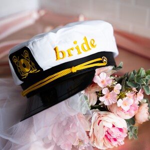 Puede incluir: Sombrero de capit&aacute;n blanco con una visera negra y ribete dorado. El sombrero tiene la palabra "bride" bordada en amarillo en la parte delantera. El sombrero est&aacute; sobre un velo blanco y un ramo de flores rosas.