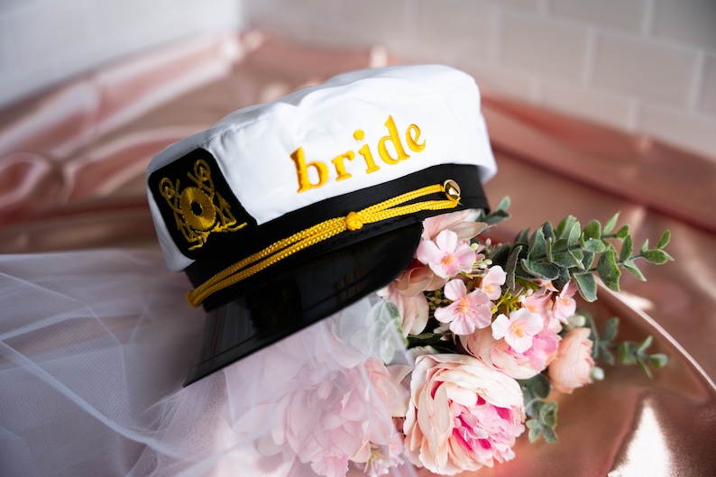 Puede incluir: Sombrero de capit&aacute;n blanco con una visera negra y ribete dorado. El sombrero tiene la palabra "bride" bordada en amarillo en la parte delantera. El sombrero est&aacute; sobre un velo blanco y un ramo de flores rosas.