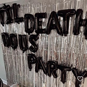May include: A silver metallic fringe curtain backdrop decorated with black letter-shaped foil balloons that spell out the phrase 'TILL DEATH DO US PARTY'. The balloons are arranged in three rows against the reflective, vertical silver streamers.