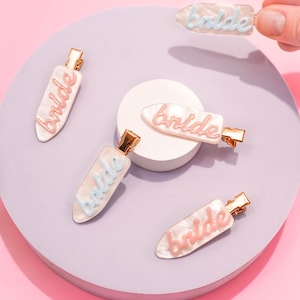 May include: Five pearlescent hair clips with the word "bride" in cursive script. The clips have gold-colored clasps and are in shades of pink and blue. The clips are arranged on a lavender surface.