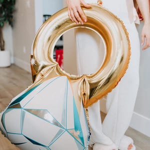 May include: A large foil balloon shaped like a diamond engagement ring. The band of the ring is metallic gold, while the diamond portion features a white and light blue geometric pattern with sharp, angular facets and small star-like accents.