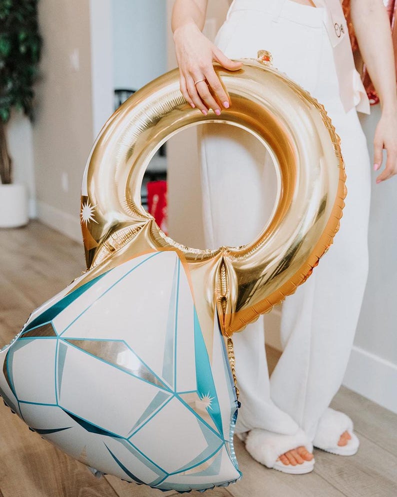 May include: A large foil balloon shaped like a diamond engagement ring. The band of the ring is metallic gold, while the diamond portion features a white and light blue geometric pattern with sharp, angular facets and small star-like accents.