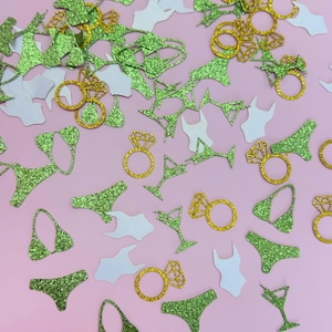 May include: A scattering of glittery green, gold, and white confetti pieces on a pink backdrop. The confetti features bikini tops and bottoms, engagement rings, martini glasses, and swimsuits. The shapes are cut out with a sparkly finish.