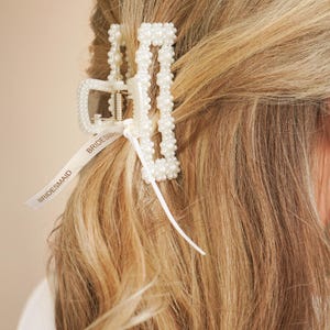 Könnte beinhalten: Eine weiße, perlenbesetzte Haarspange mit einem Band mit der Aufschrift "Bridesmaid" ist in blondem Haar platziert. Die Spange besteht aus weißem Kunststoff und hat ein Metallscharnier.