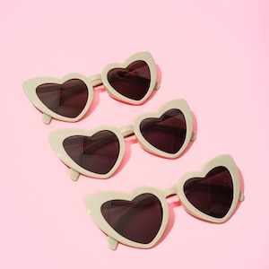 May include: Four pairs of heart-shaped sunglasses with dark lenses and a light green frame are arranged on a pink background. The sunglasses have a retro, playful design, perfect for adding a touch of fun to any outfit.