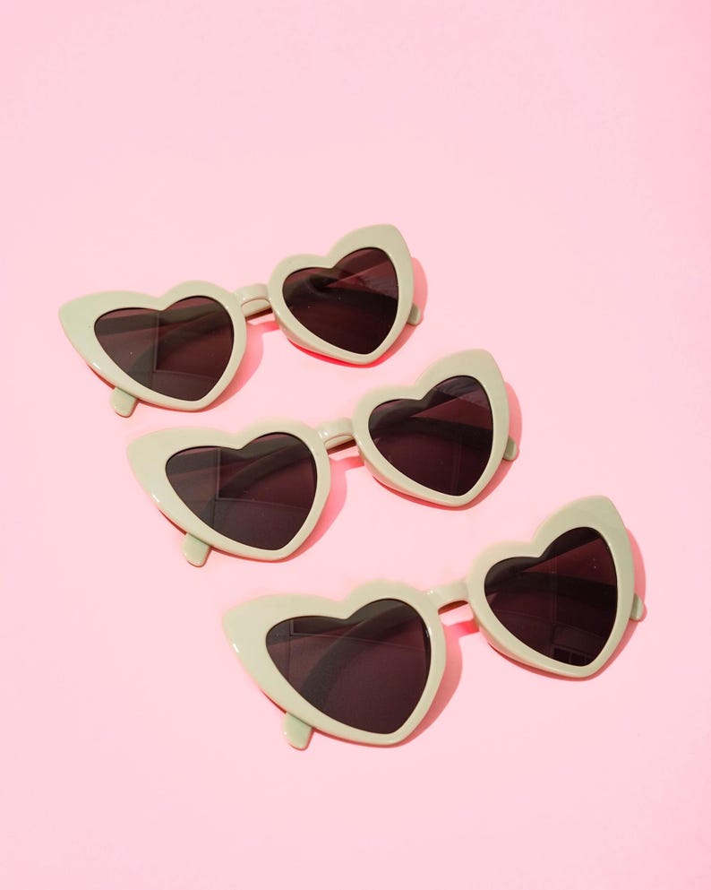 May include: Four pairs of heart-shaped sunglasses with dark lenses and a light green frame are arranged on a pink background. The sunglasses have a retro, playful design, perfect for adding a touch of fun to any outfit.