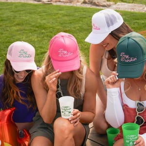Puede incluir: Cuatro gorras de camionero en rosa, blanco y verde con el texto "Day Drinkin" bordado en la parte delantera. Las gorras tienen una parte trasera de malla y una visera curva. La imagen muestra las gorras siendo usadas por personas.