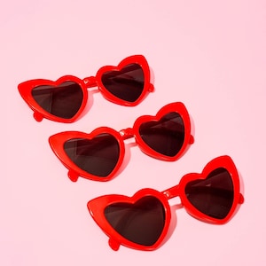 May include: Four pairs of red heart-shaped sunglasses with dark lenses are arranged diagonally on a pink background. The sunglasses have a bold, retro style, perfect for a fun, fashionable look. The frames are a vibrant red.