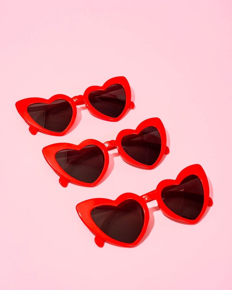 May include: Four pairs of red heart-shaped sunglasses with dark lenses are arranged diagonally on a pink background. The sunglasses have a bold, retro style, perfect for a fun, fashionable look. The frames are a vibrant red.