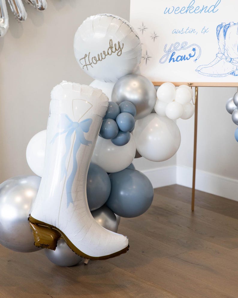 May include: A festive balloon arrangement featuring a white cowboy boot, silver, white, and blue balloons. A round balloon says "Howdy" in gold script. A sign reads "weekend Austin, TX Yee Haw".